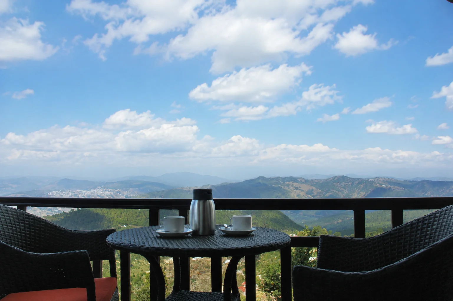 Balcony table set with two cups and a flask, with wicker chairs and a wide view of green mountains under a blue sky.