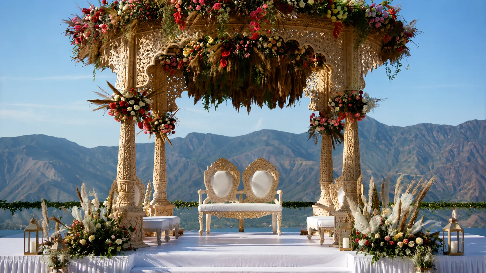 Carved wooden mandap with lush floral canopy, two ornate chairs, and mountain lake backdrop.