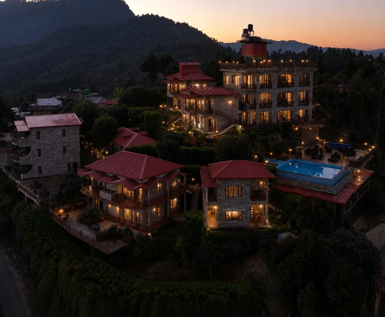 Hillside resort with red-roofed stone buildings and a lit pool, glowing at dusk against forested mountains.