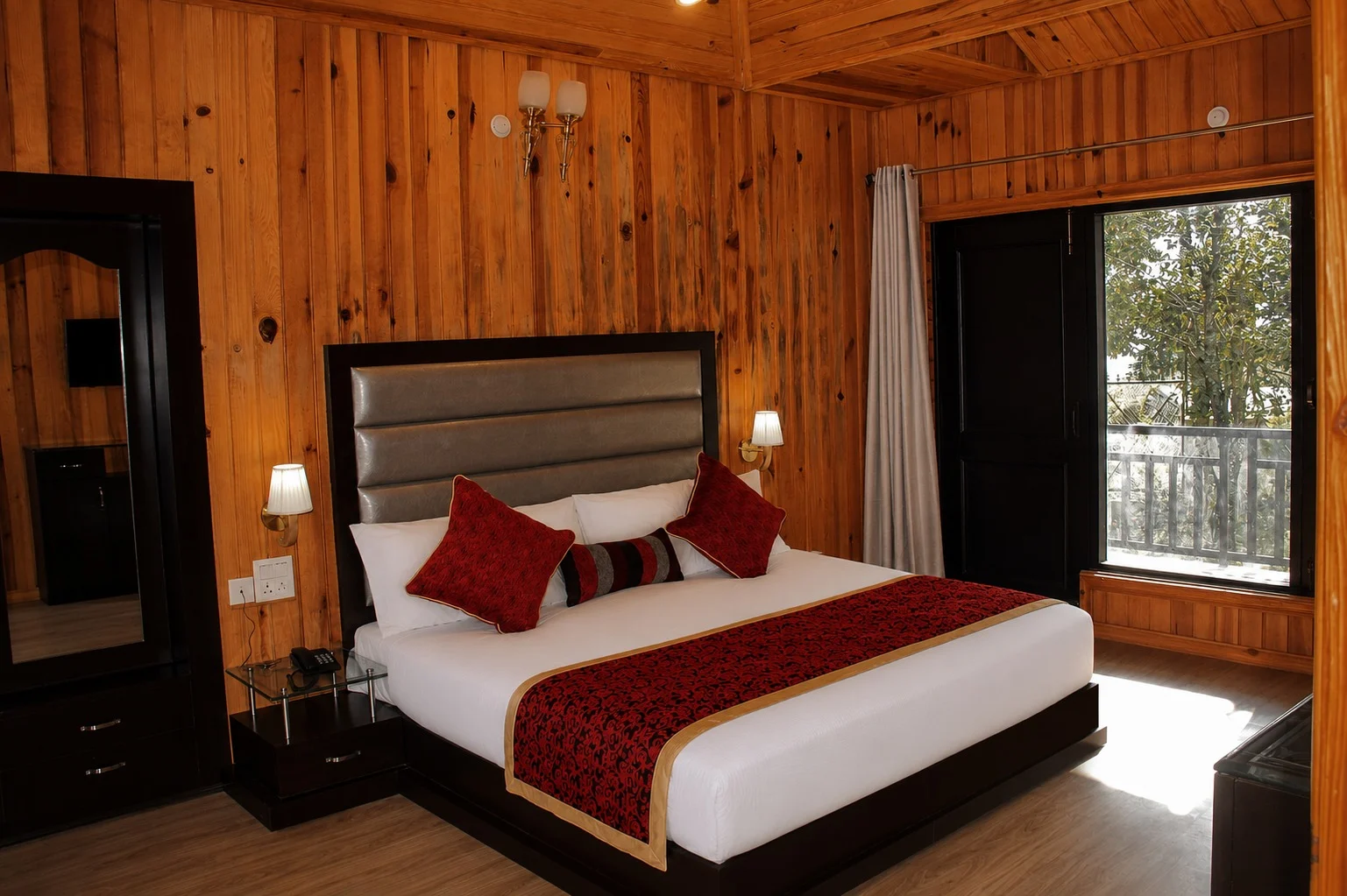 Wooden-panelled hotel room with a king bed, red cushions and a balcony view.