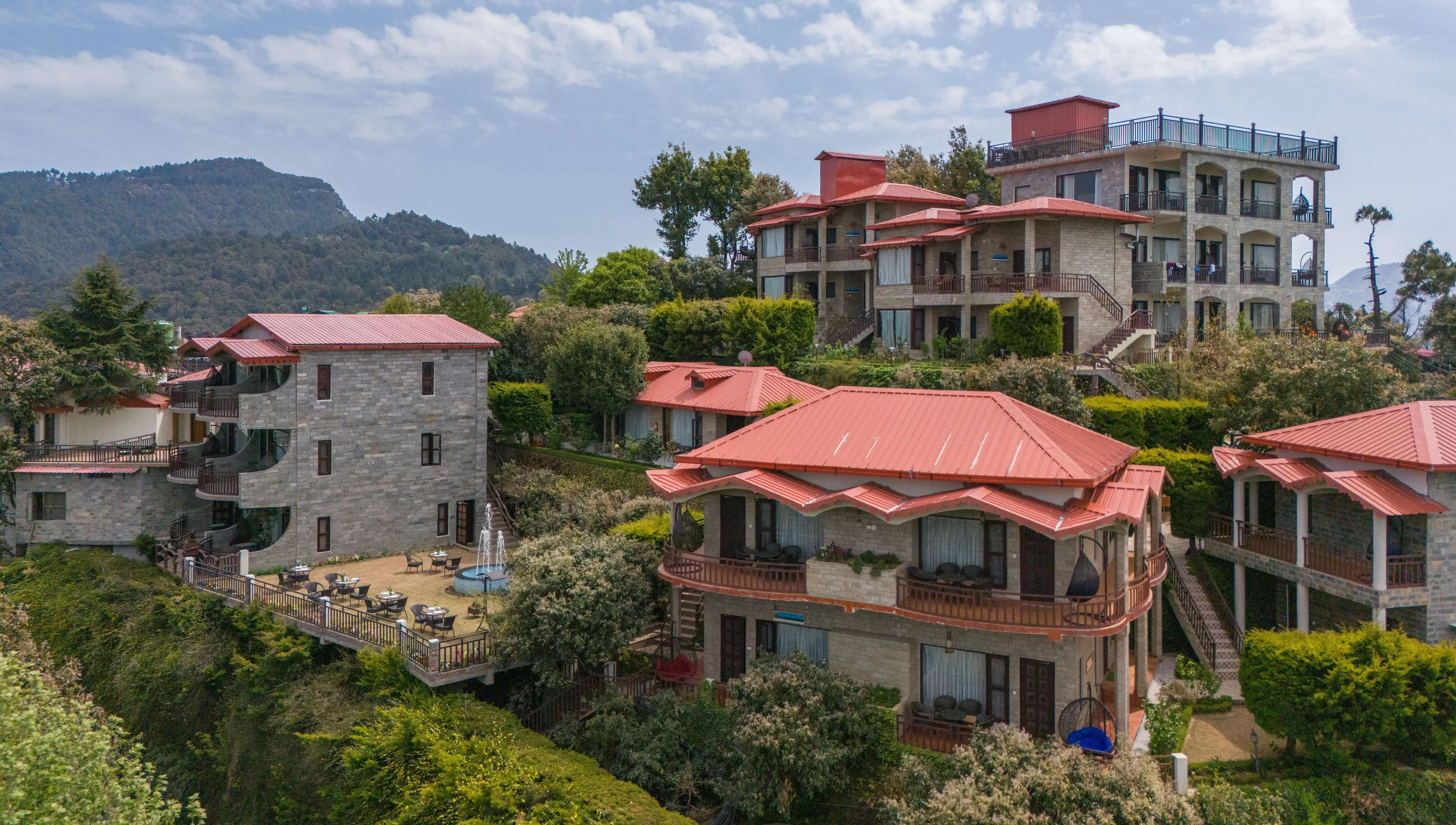 Aerial view of a sprawling luxury resort with several red-roofed stone cottages nestled on a green hillside in the Himalayas.