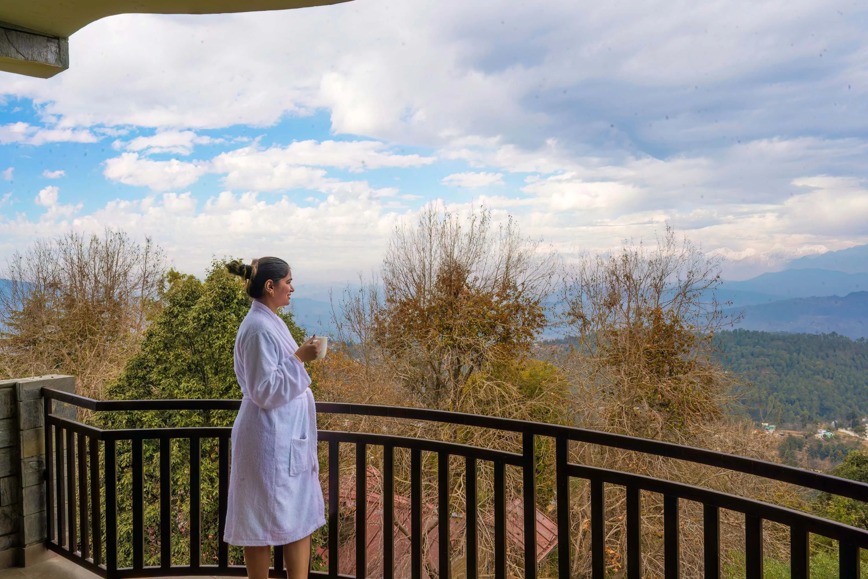Woman enjoying a morning cup of tea on a mountain-view balcony, waking up above the clouds amidst serene Himalayan landscapes.